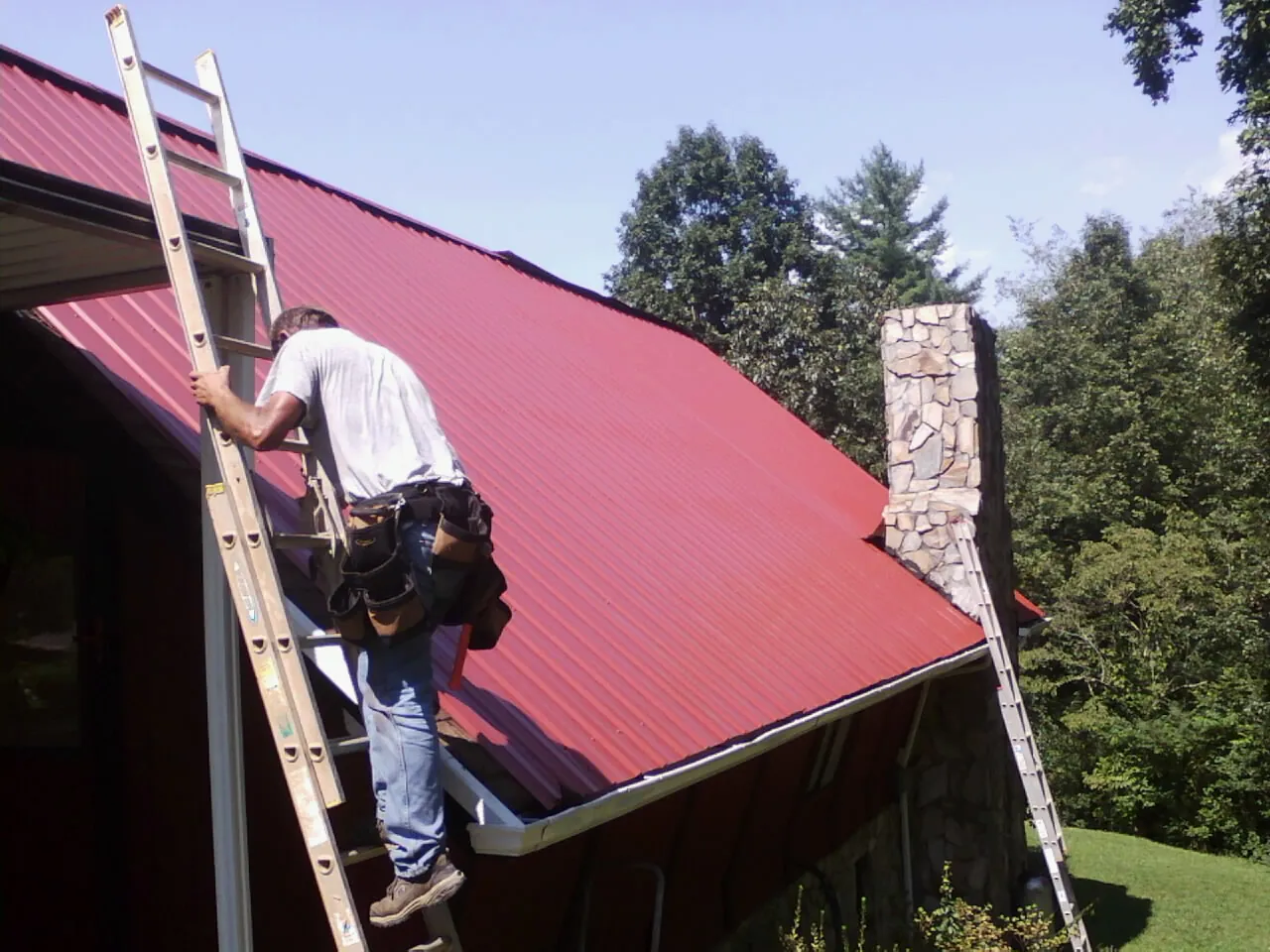 Roofer on a ladder beside a red metal roof and stone chimney — Soffit & Fascia Repair by Roofing Brothers in Bucoda